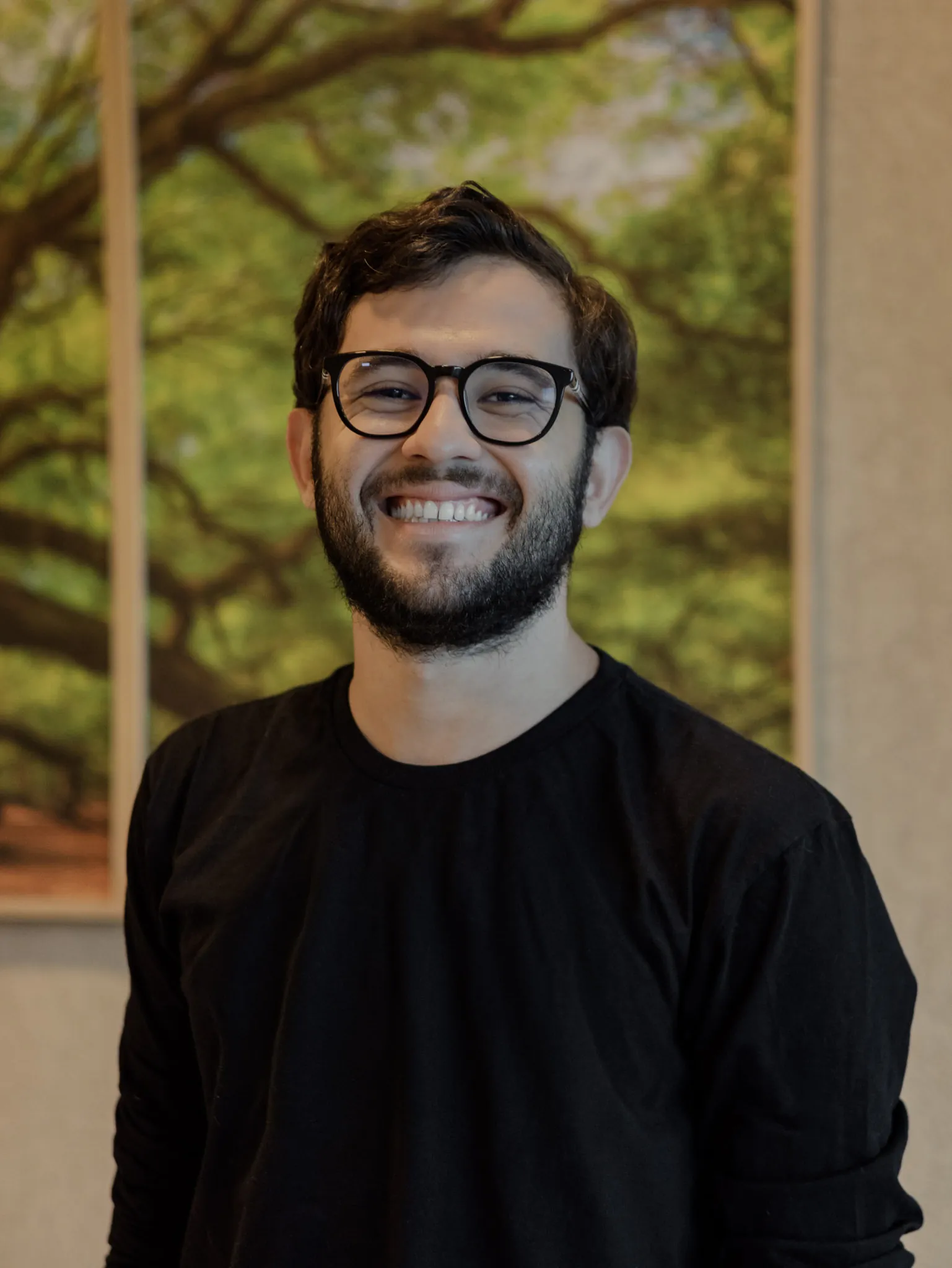 Retrato de Breno Gouveia. Homem de óculos e barba sorri para a câmera, vestindo camiseta preta, diante de uma parede com uma pintura desfocada de uma enorme árvore em tons de verde e marrom ao fundo.